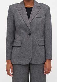 Grå texturerad blazer med en svart knapp, slagkrage, två framfickor och skräddarsydd passform. Matchas med en svart topp under.