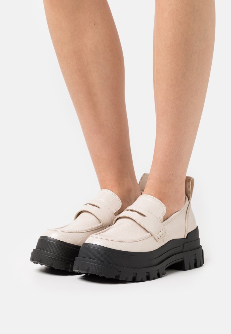 Mocassins couleur crème avec motif texturé ressemblant à du crocodile, semelle noire épaisse, accents à deux sangles, et une coupe basse.