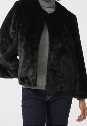 Sort jakke i imiteret pels, båret over en grå rullekravetrøje og mørkeblå jeans, vist fra hals til hofter.