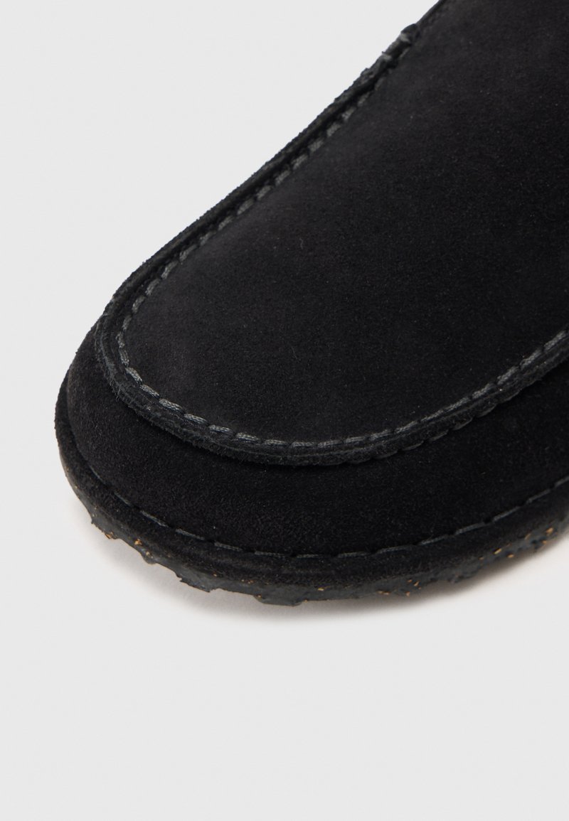 Primer plano de un zapato mocasín de ante negro, con costuras en gris que contrastan y una textura suave, acompañado de una suela de goma flexible.