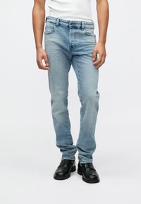 Jeans de mezclilla azul claro con corte slim, cinco bolsillos, pasadores para cinturón y un ligero desgaste. Llevados con zapatos negros tipo mocasín.