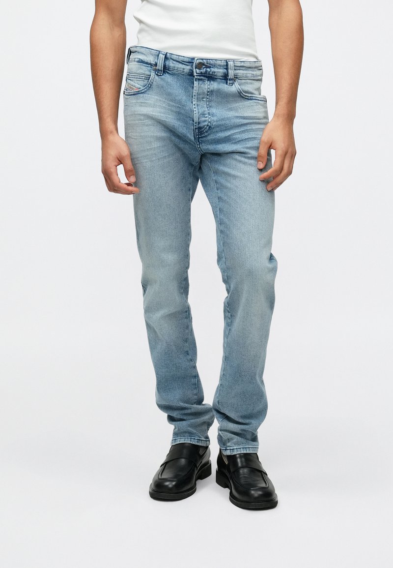 Jeans de mezclilla azul claro con corte slim, cinco bolsillos, pasadores para cinturón y un ligero desgaste. Llevados con zapatos negros tipo mocasín.