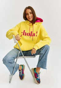 Žltý mikina s ružovými šnúrkami a textom "¡hola!" na prednej strane, skombinovaná so svetlými, mierne ošúchanými džínsami a farebnými teniskami.