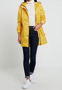 Geel waterdichte jas met capuchon, elastische manchetten en drukknoopsluiting, gedragen over een witte coltrui en donkere skinny jeans. Witte sneakers.