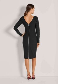 Frau in einem figurbetonten schwarzen Kleid mit tiefem V-Ausschnitt und einem langen silbernen Reißverschluss, kombiniert mit roten hochhackigen Sandalen, steht vor einer schlichten Wand.