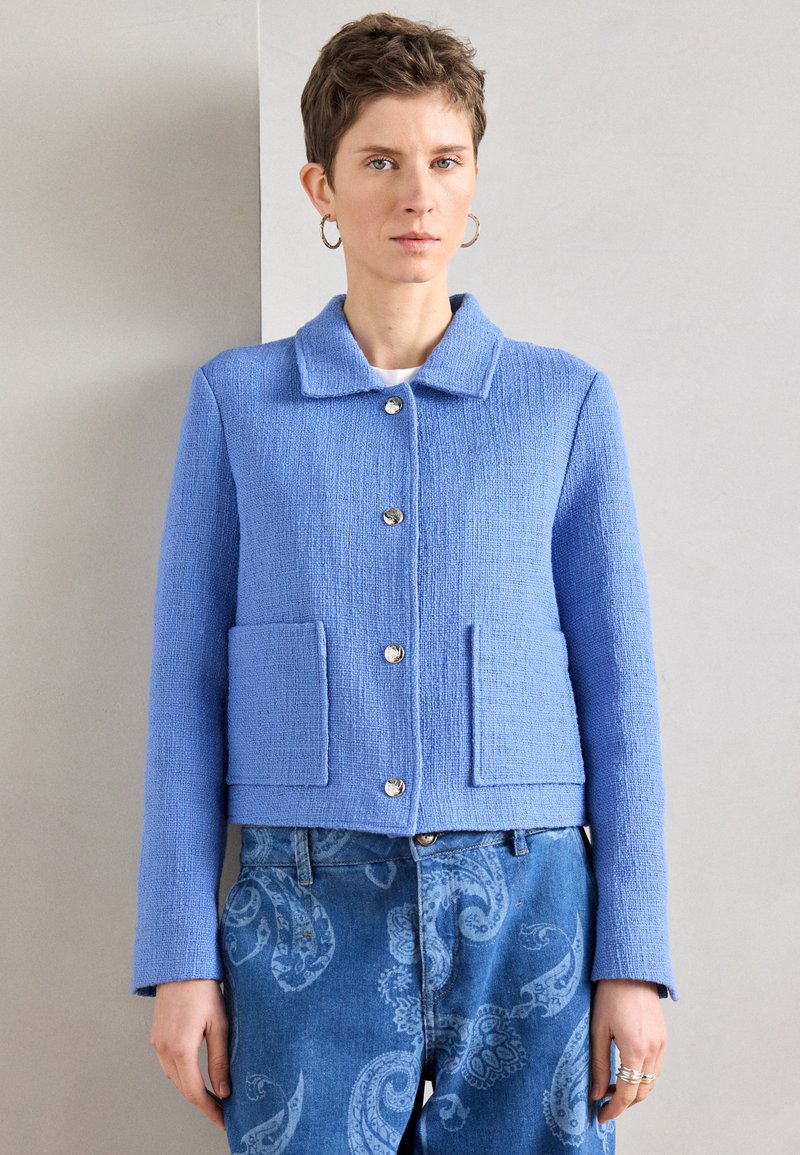 Μπλε υφασμάτινο μπουφάν με γιακά, δύο μπροστινές τσέπες και κλείσιμο με γυαλιστερά ασημένια κουμπιά. Φορεμένο πάνω από μπλε τζιν με σχέδια.