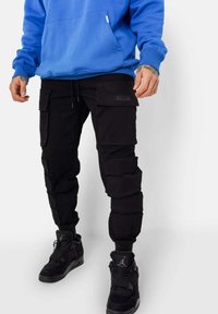 Zwarte cargobroek met meerdere zakken, gecombineerd met een blauwe hoodie. De stof lijkt textuur te hebben, met elastische boorden en een relaxte pasvorm.