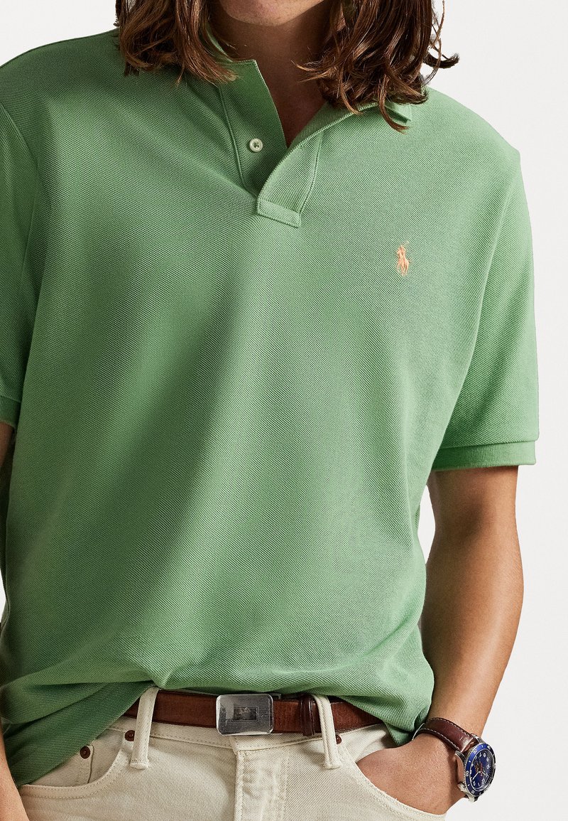 Groene polo met een gestructureerde stof, voorzien van een placket met twee knopen en een geborduurd logo op de borst. Draagt met een bruine riem.