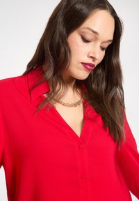 Czerwona bluzka na guziki z głębokim dekoltem w kształcie litery V, długimi rękawami i gładką fakturą. Charakteryzuje się luźnym krojem i widocznym złotym łańcuszkiem.