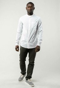 Weißes kurzärmeliges Hemd mit Brusttasche, kombiniert mit dunkelgrauen Hosen und grauen Sneakers. Einfaches Design, glatte Stofftextur.