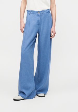 Persoon met lichtblauwe wijde spijkerbroeken en witte schoenen, staand tegen een effen lichte achtergrond.