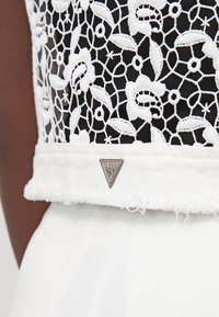 Biała koronkowa bluzka z kwiatowym wzorem, na czarnym tle, z postrzępionym dołem. Na dole znajduje się srebrne trójkątne logo "Guess".