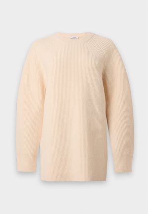 Femme aux cheveux noirs portant un pull beige côtelé, un jean large foncé et des ballerines noires, debout devant un fond blanc uni.