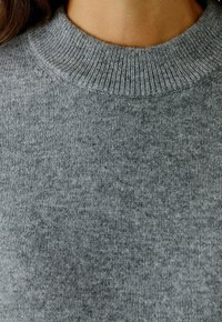 Nahaufnahme einer Person, die einen grauen gerippten Rundhals-Pullover trägt, wobei der Hals- und Oberbrustbereich zu sehen ist.