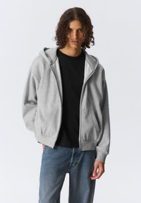 Sweat à capuche gris avec une fermeture éclair frontale, des poches spacieuses et des poignets côtelés. Porté sur un t-shirt noir et associé à un jean bleu.