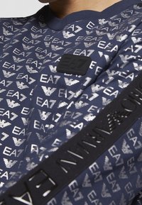 Sudadera azul marino con un patrón repetido del logo plateado de EA7, cuello acanalado y un parche de logo de goma negra en el pecho. Detalle de tejido texturizado.