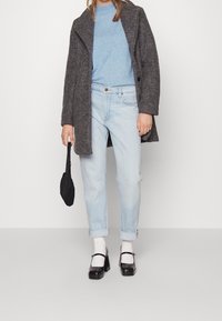 Manteau en laine gris, pull bleu clair, jean en denim délavé clair, chaussures à plateforme noires, chaussettes blanches, tenant un petit parapluie noir.