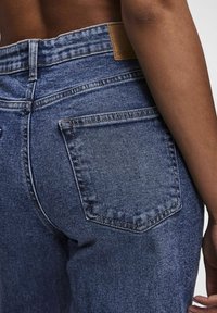 Mörkblå denimjeans med hög midja, bakfickor och en läderpatch på midjebandet. Texturerad tyg med synlig sömnad.