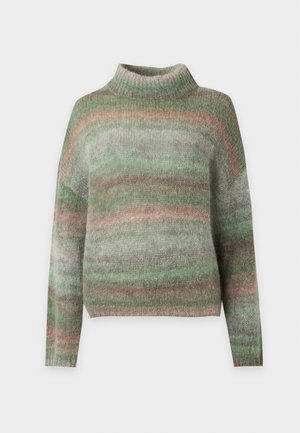 Personne portant un pull ample rayé vert et beige et un pantalon large sombre surdimensionné avec des chaussures marron, debout devant un fond uni.