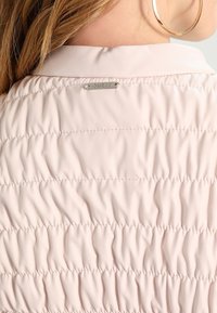 Giacca trapuntata rosa chiaro con colletto liscio e una etichetta argentata con logo sul retro. Superficie texturizzata con dettagli arricciati.