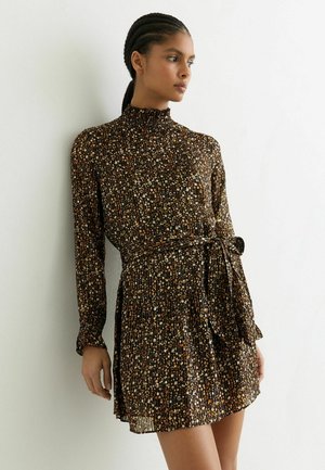 Kvinde med flettet hår iført en langærmet, højhalset blomsterkjole med taljebånd, stående mod en hvid væg og kiggende til siden.