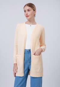 Cardigã de cor pêssego, feito de um material de malha suave, design com frente aberta, punhos canelados e dois bolsos frontais, combinado com jeans de um tom azul claro.