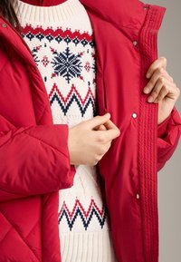 Rød polstret jakke med quiltet struktur, lukkes med trykknapper, lagdelt over en cremefarvet strikket sweater med snefnug- og zigzag-mønster.