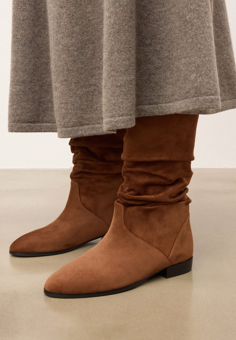 Bottines en daim marron avec un design loose, bout pointu et petit talon noir, associées à une jupe grise. Texture lisse dans l'ensemble.