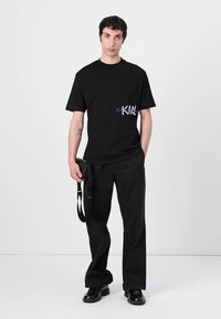 Fekete pamut póló rövid ujjakkal, a bal mellen egy színes "KARL" logóval. Fekete, bő szárú nadrággal viselve.