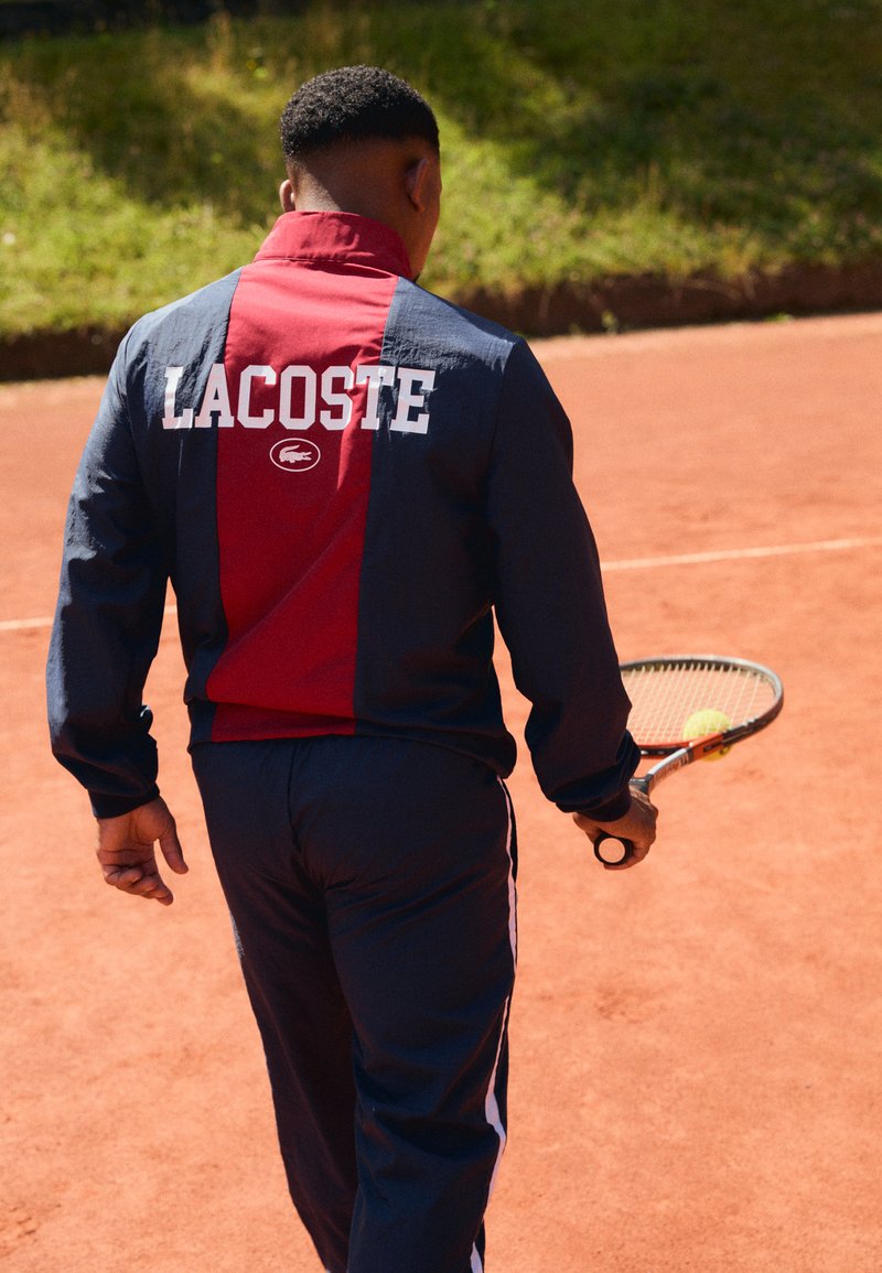 Survêtement Lacoste en bleu marine et bordeaux, avec le logo bien en évidence au dos. Comprend des accents blancs. Raquette de tennis à la main, de dos.