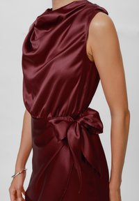Burgundy satin dress with a draped neckline, an elastic waist, and a large bow accent at the side. Smooth texture, sleeveless design.