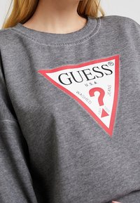 Harmaa huppari, joka on valmistettu puuvillasekoitteesta, jossa on suuri punavalkoinen kolmiomainen logo ja teksti "GUESS U.S.A. WASHED JEANS" sekä kysymysmerkki.