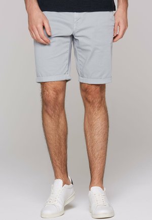 Man wearing light gray rolled-hem shorts, white sneakers, and a dark top, standing against a plain gray background.