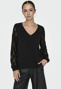Czarny top z dekoltem w serek i długimi koronkowymi rękawami. Dół jest wykonany z jednolitego materiału, a rękawy mają kwiatowy wzór z koronki. Połączony z czarnymi spodniami.