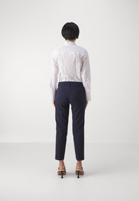 Chemise blanche boutonnée, pantalon sur mesure bleu marine et sandales noires à talons hauts. Tissu lisse, coupe ajustée, vue de dos.