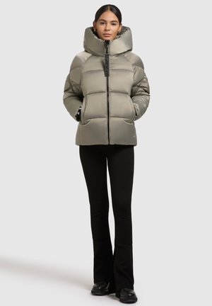 Chaqueta de plumas verde oliva con un cuello alto, cierre de cremallera frontal y grandes secciones acolchadas. Combinada con pantalones de campana negros y botas.