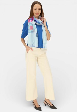 Kobieta ubrana w niebieski sweter z krótkim rękawem, jasnoniebieski kwiecisty szal, kremowe szerokie spodnie oraz czarne czółenka na niskim obcasie typu slingback, stoi z ręką w kieszeni.