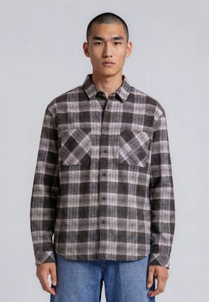 Jeune homme aux cheveux courts portant une chemise à carreaux grise et noire boutonnée et un jean bleu, debout devant un fond clair uni.