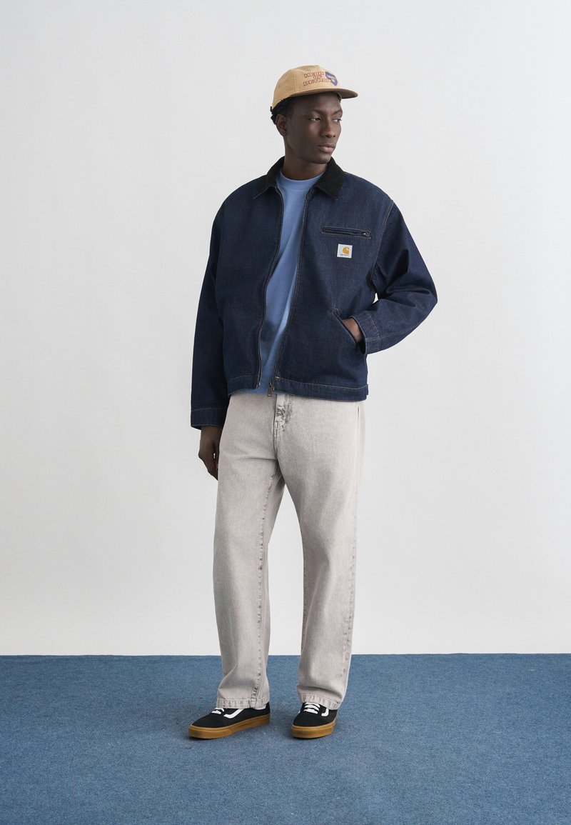 Hombre con gorra beige, chaqueta azul oscuro, camisa azul clara, vaqueros gris claro y zapatillas negras de pie sobre una alfombra azul frente a una pared blanca.