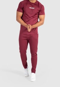 Bordeaux tränings t-shirt och byxor set, med rundad hals, slim fit och mjuk textur. Vit logotyp visas på t-shirten. Vita sneakers.