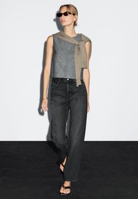 Graues ärmelloses Top mit einem beigen Bindedetail, kombiniert mit hochgeschnittenen schwarzen Jeans mit geradem Schnitt. Schwarze Sandalen runden den Look ab.