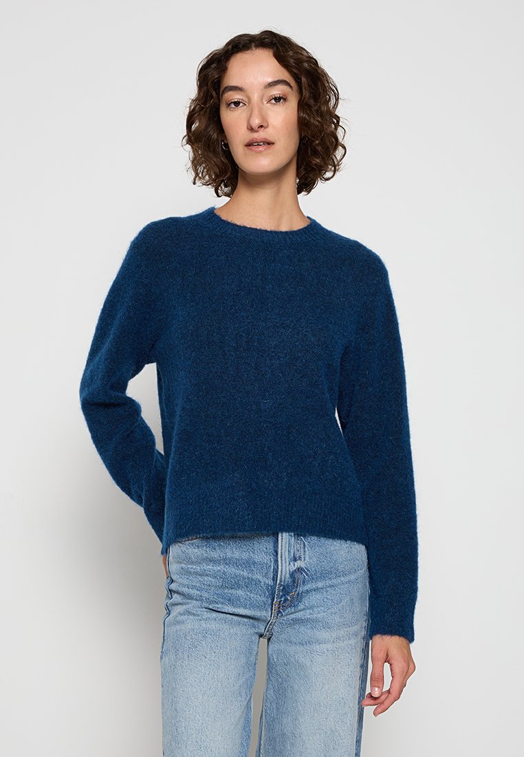 Kvinna med lockigt hår som bär en blå fluffig tröja och ljusblå jeans, stående mot en enkel ljus bakgrund.