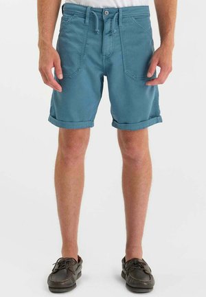 Man wearing teal blue rolled-hem shorts with front pockets and dark brown casual lace-up shoes, standing against a plain background.