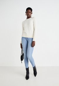 Pull beige à manches longues, texture douce, et accents de boutons aux poignets. Porté avec un jean skinny bleu clair et des bottes noires à bout pointu.