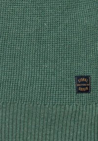 Pulover tricotat de culoare verde închis, cu textură cu nervuri. Prezintă o etichetă maro pe care este imprimat "SIGNAL" și "QUALITY SINCE 1971" în aur.