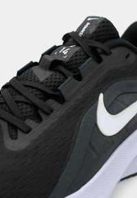 Μαύρο παπούτσι για τρέξιμο Nike με λευκό λογότυπο Swoosh, πλέγμα στο πάνω μέρος και μαύρα κορδόνια, που δείχνει λεπτομέρεια από τη γλώσσα με μέγεθος και το κείμενο "Downsh."