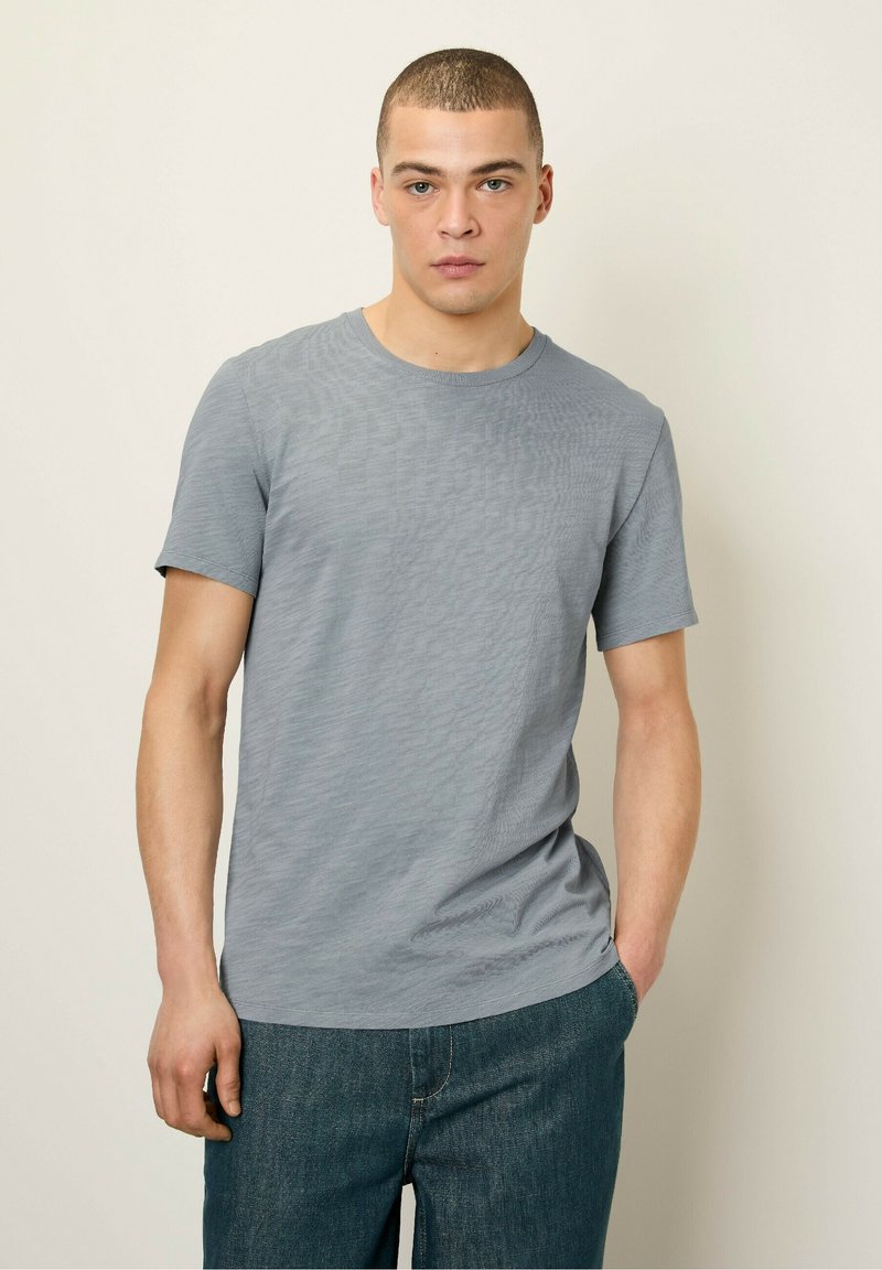 Pelēks īsām piedurknēm T-krekls ar smalku strukturētu dizainu, apaļu kakla izgriezumu un brīvu piegriezumu, kopā ar tumši ziliem džinsiem.