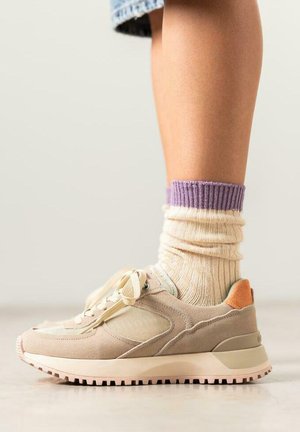 Chaussure de sport beige avec un dessus en daim et en maille, semelle en caoutchouc rose et accent orange au talon. Associée à des chaussettes crème et violettes.