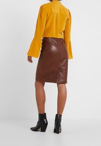 Blusa gialla a maniche lunghe con maniche a campana abbinata a una gonna di pelle marrone aderente. Stivaletti neri alla caviglia con tacco a blocco e dettaglio con zip visibile.