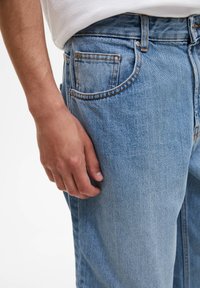 Mužská ruka spočívající vedle kapsy a stehna světle modr�ých džínů, oblečená v bílém tričku.